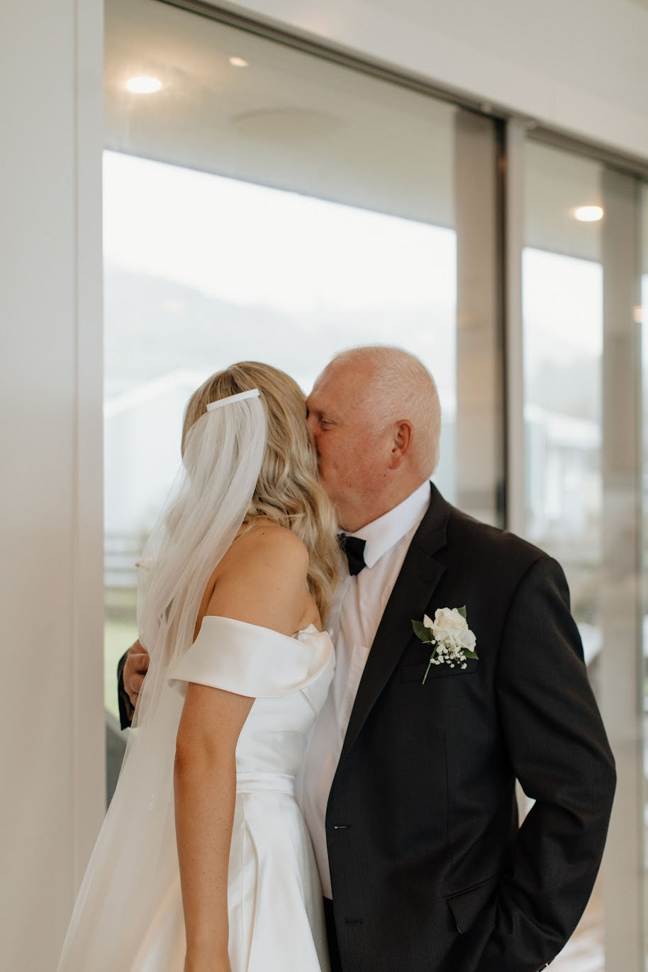 Man and woman in wedding attire standing close together in front of a reflective surface wearing a buttonhole