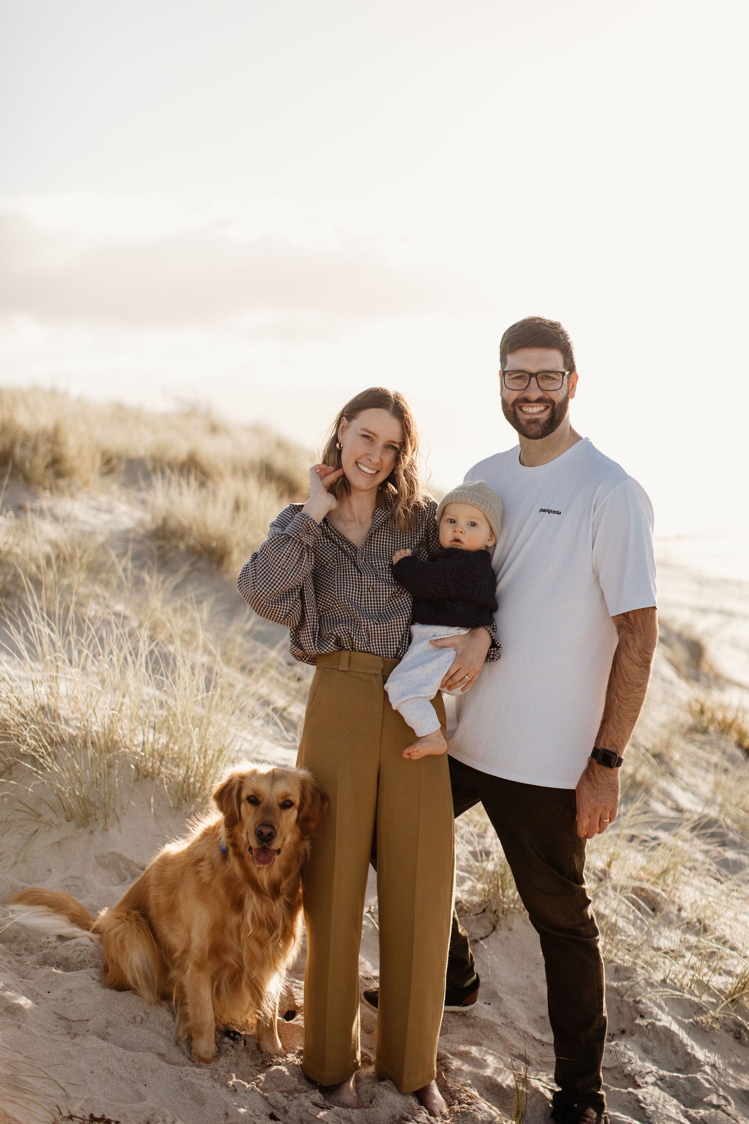 Family of three with a dog standing on a sandy beach.