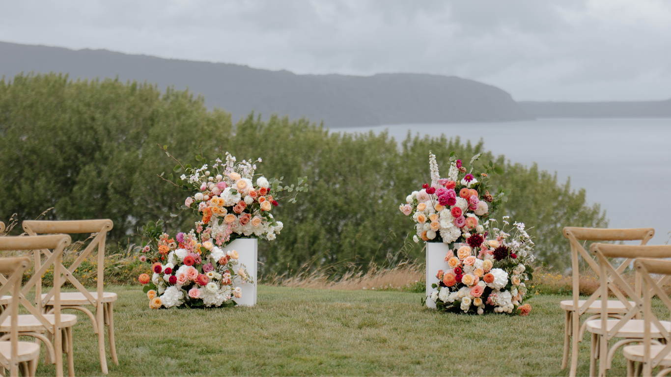 Floral plinth arrangements on a grassy area with chairs and a scenic background