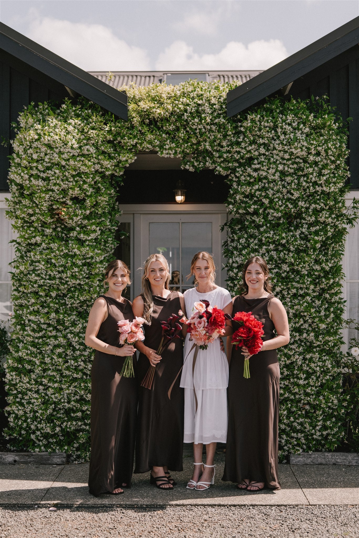 Four women standing in front of a building with greenery, holding bouquets.