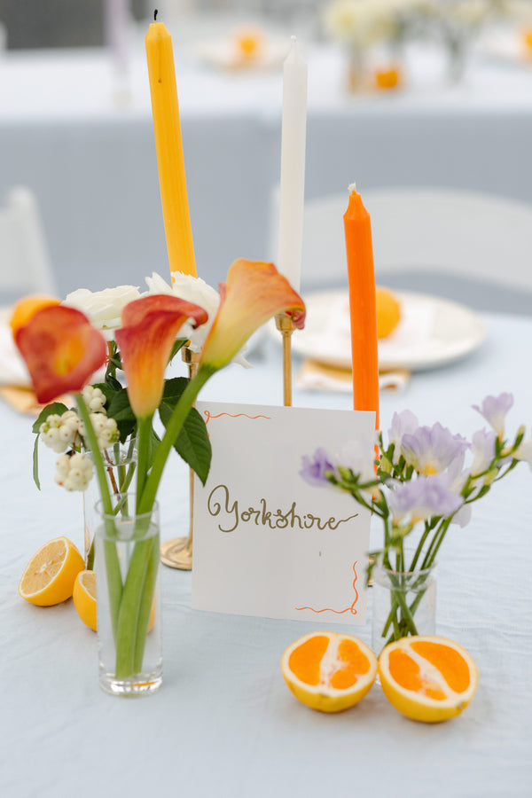 Decorative table setting with flowers, candles, and a card on a light surface.