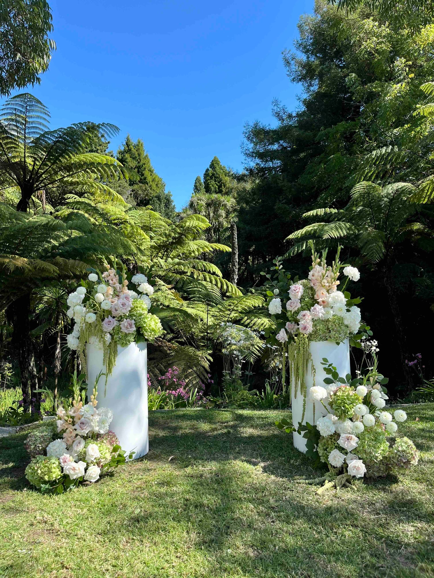 Floral Plinth arrangements in a garden setting with ferns and blue sky.