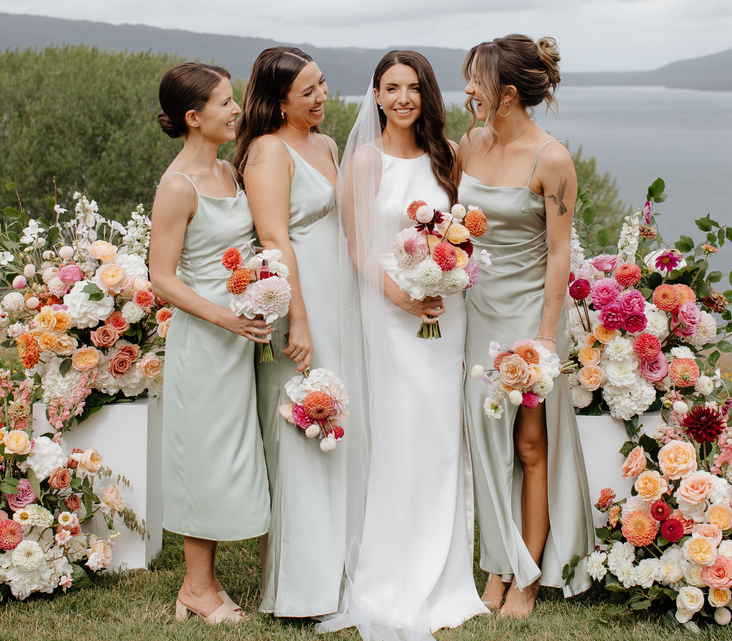 Bride with three bridesmaids standing in front of floral plinth decorations by a lake.