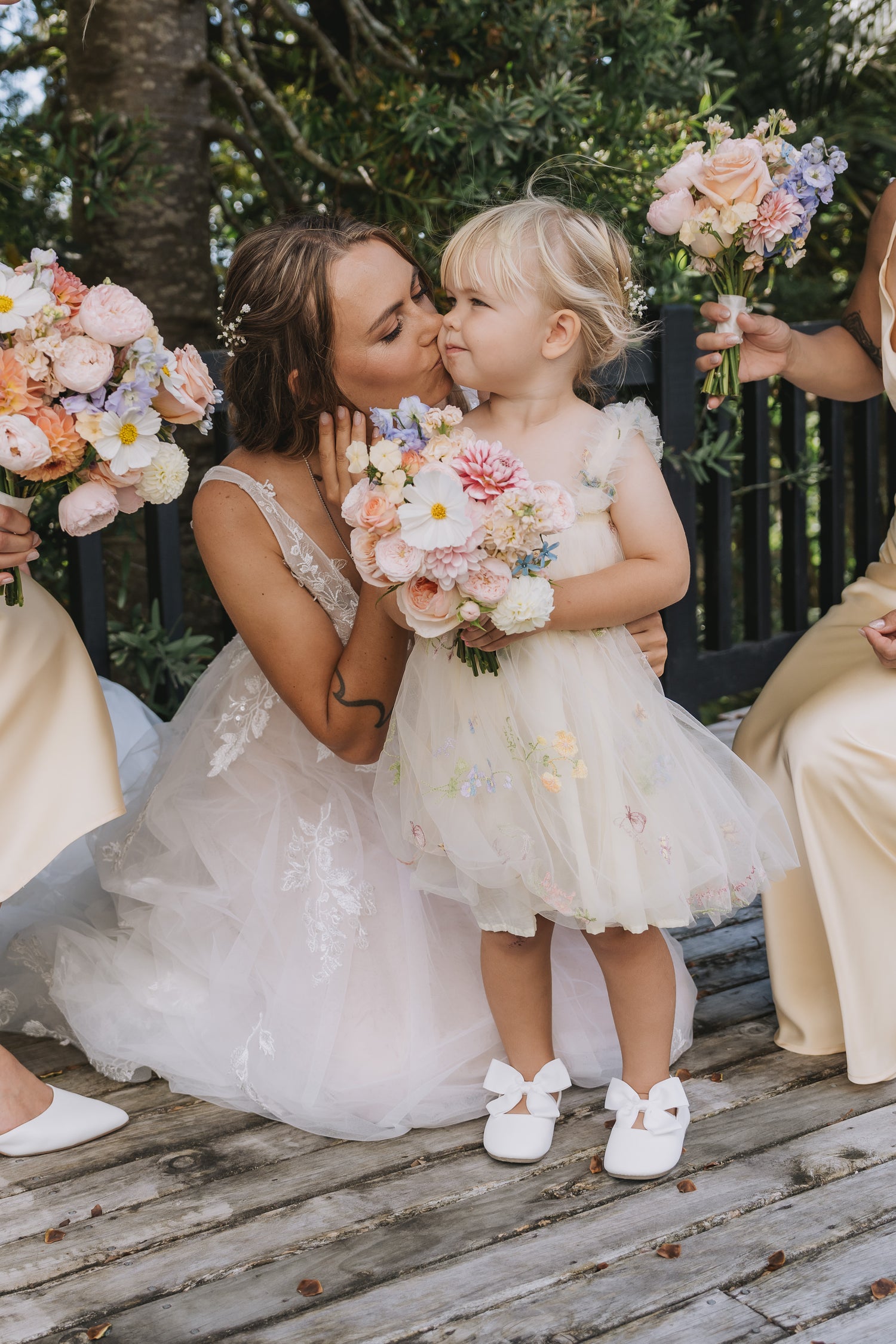 Woman in a wedding dress kissing a young girl in a floral dress on a wooden deck with flowers in the background.