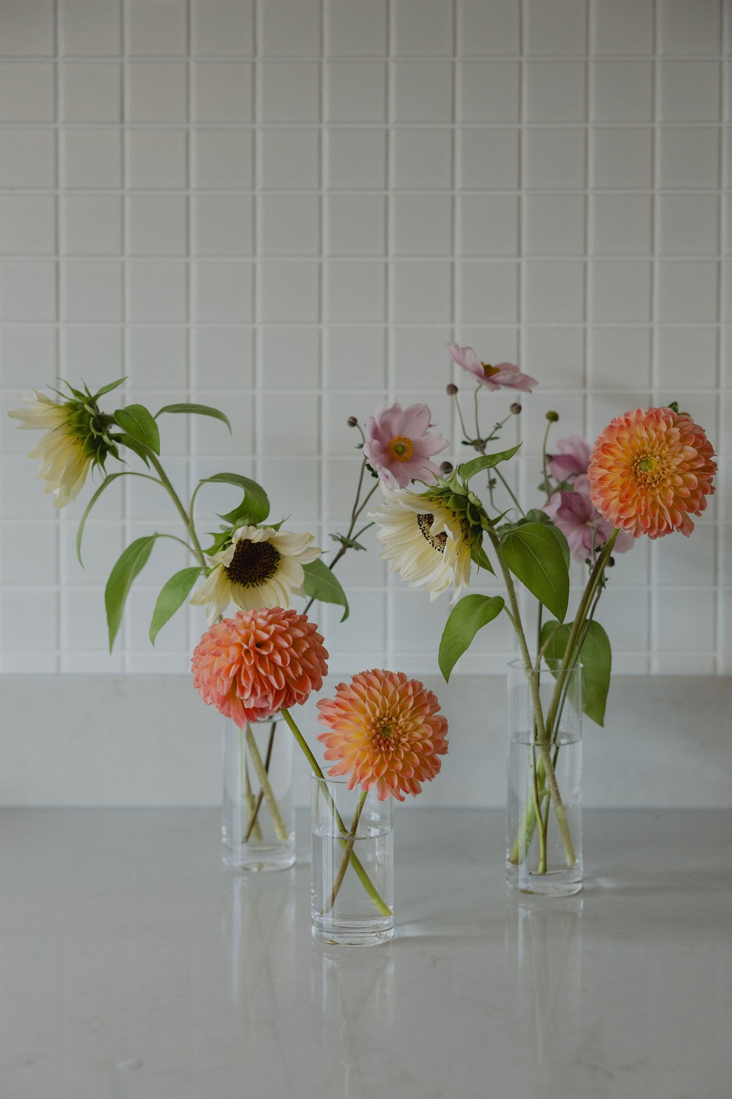 Three glass vases with pastel flowers on a reflective surface and tiled wall background