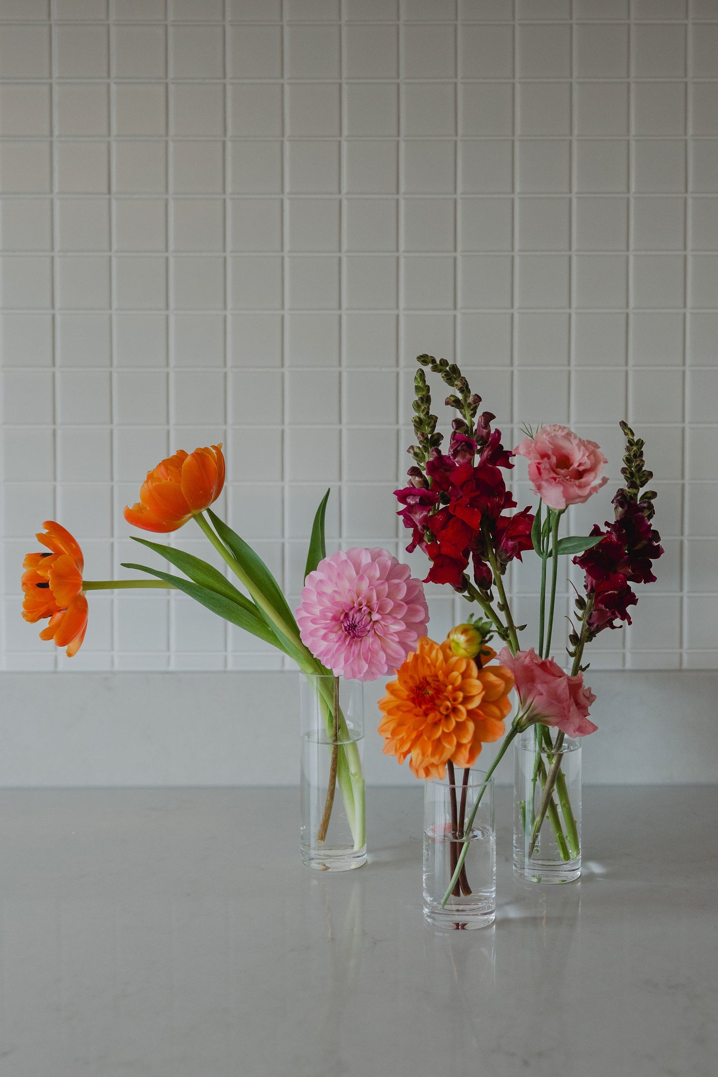 Three glass vases with colourful flowers on a white surface and tiled wall background