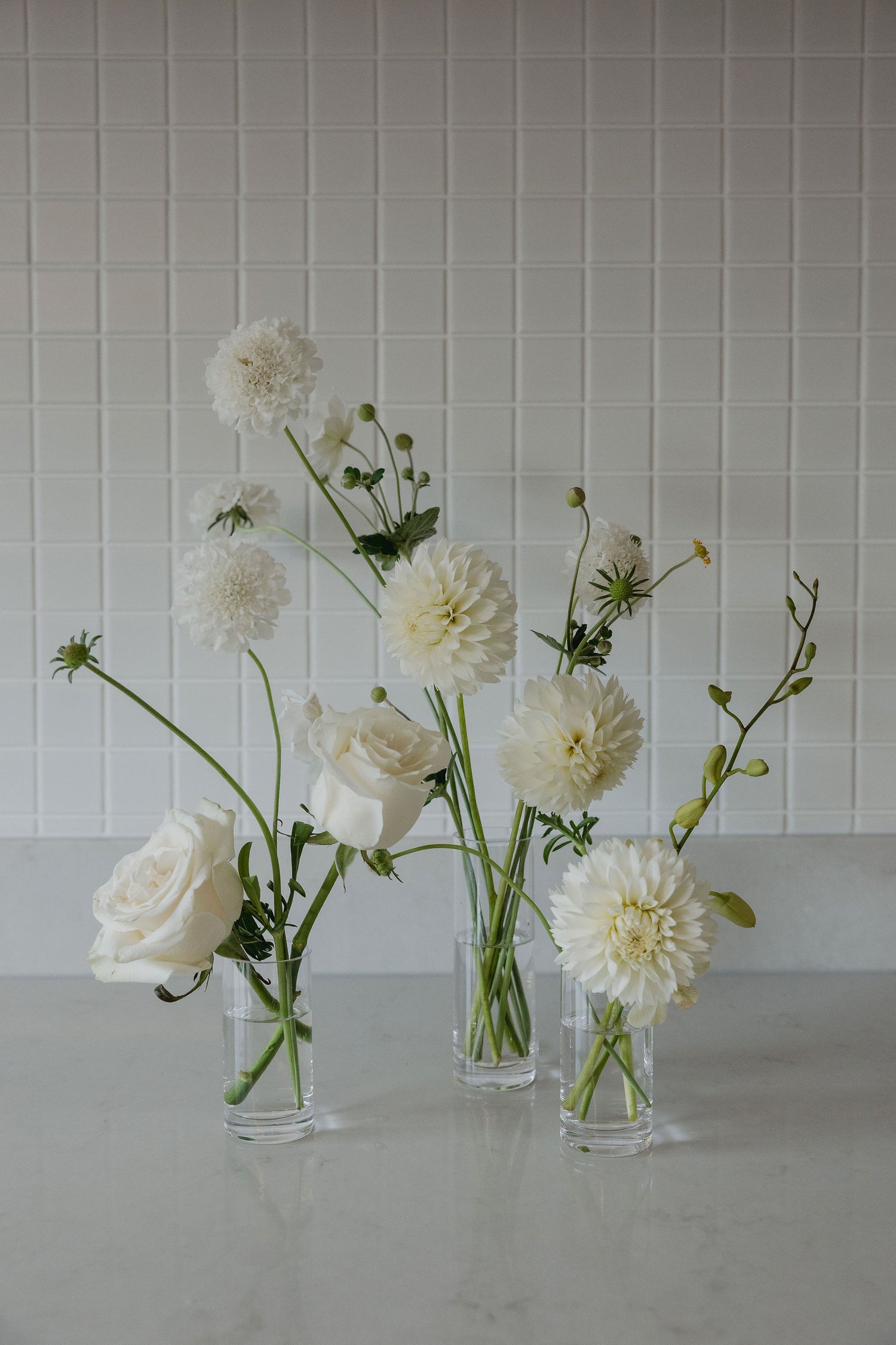 Three clear glass vases with white flowers on a light gray surface against a white tiled wall.
