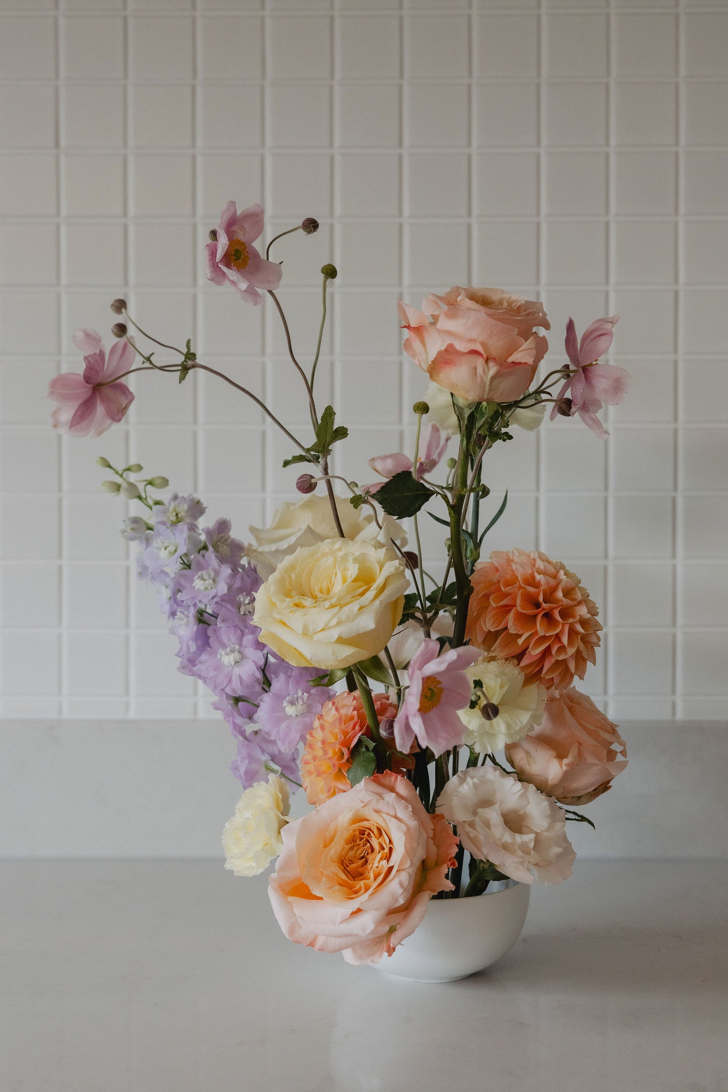 Pastel floral arrangement in a white bowl against a white tiled wall.