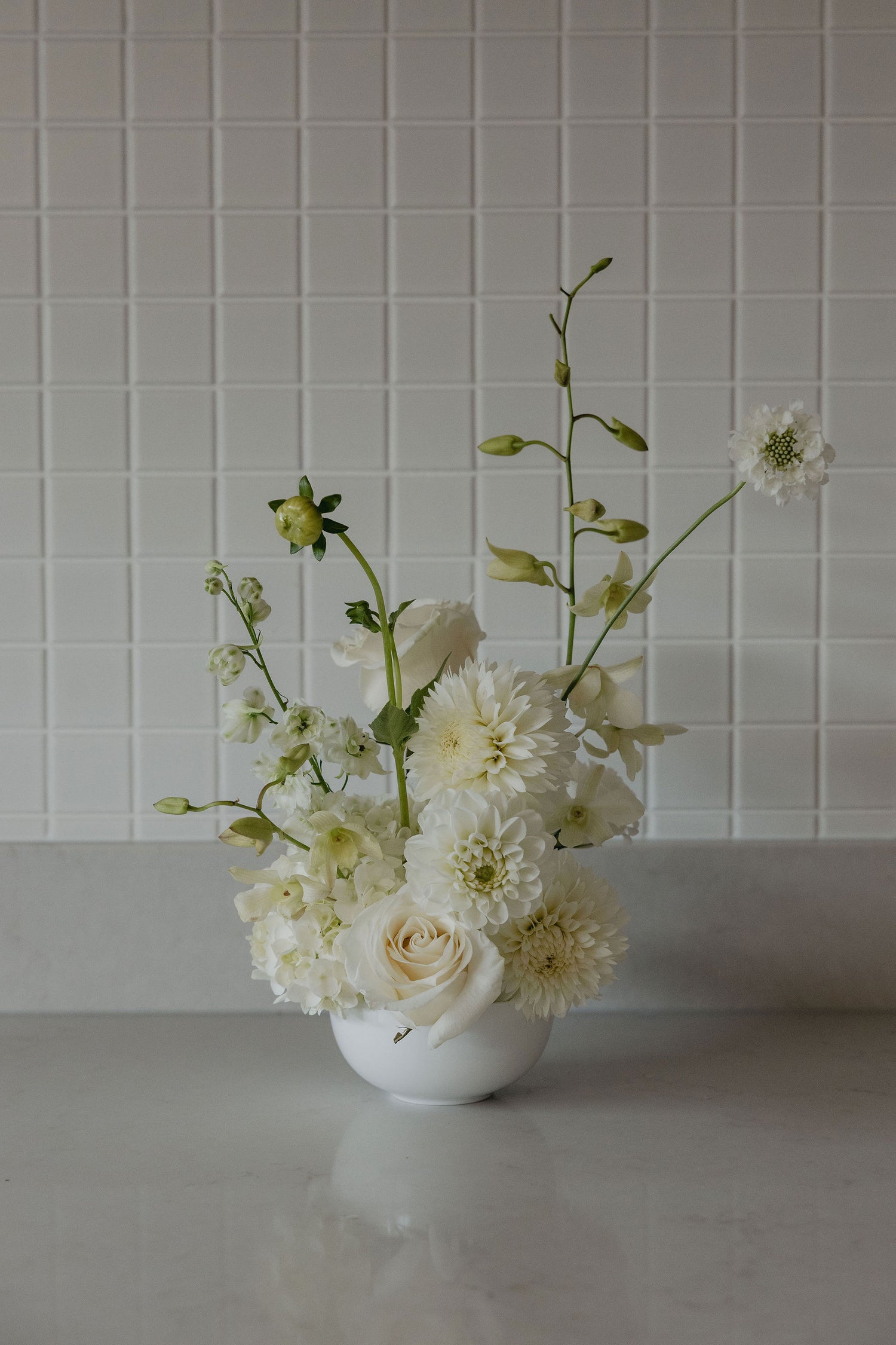 White floral arrangement in a vase against a white tiled wall.
