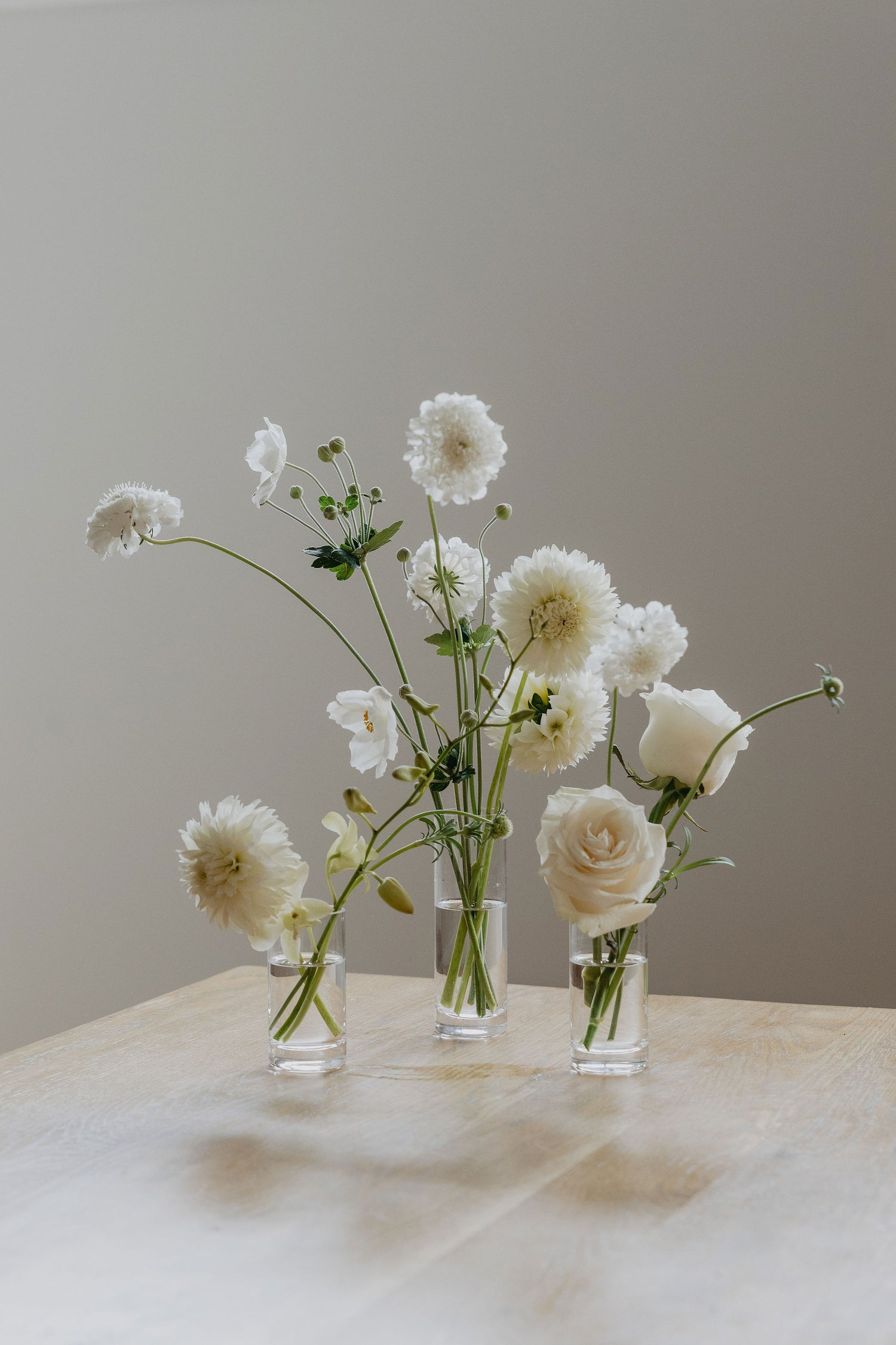 Three small glass vases with white flowers on a marble surface against a plain background