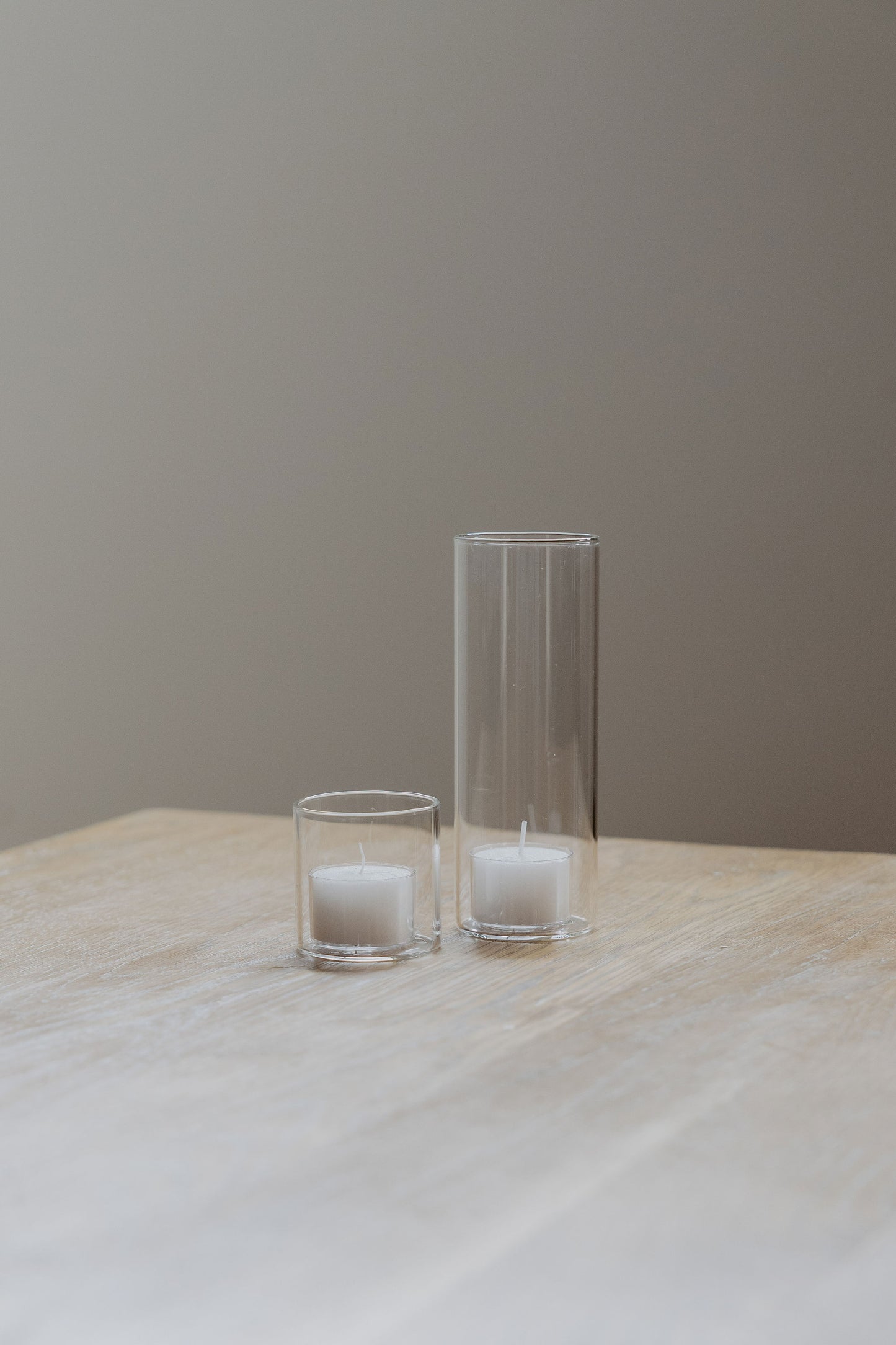 Two glass tealight candle holders with candles on a wooden table against a plain wall.