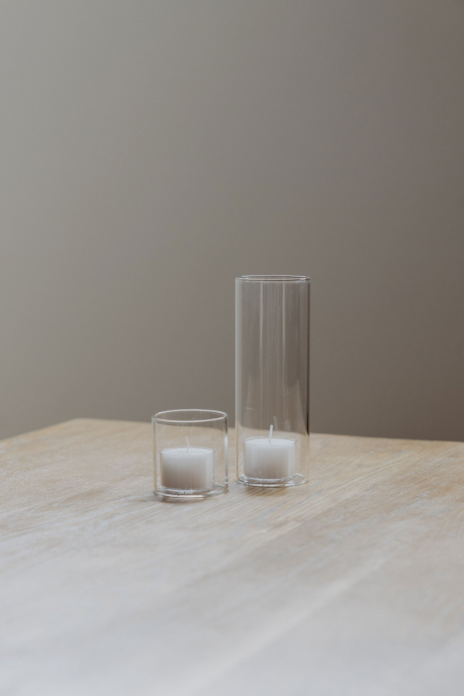 Two glass tealight candle holders with candles on a wooden table against a plain wall.