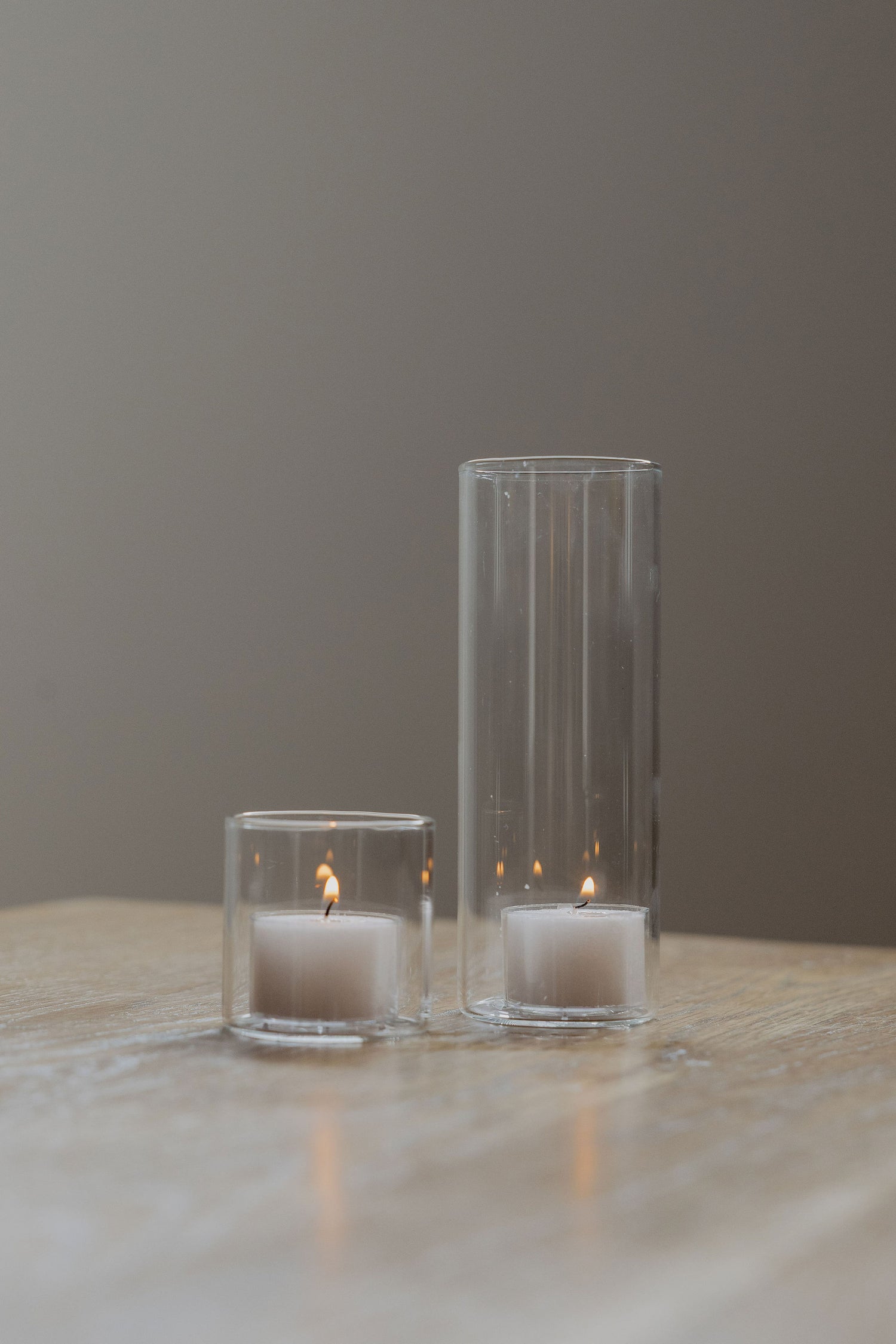 Two clear glass candle holders with candles on a wooden surface against a gray background