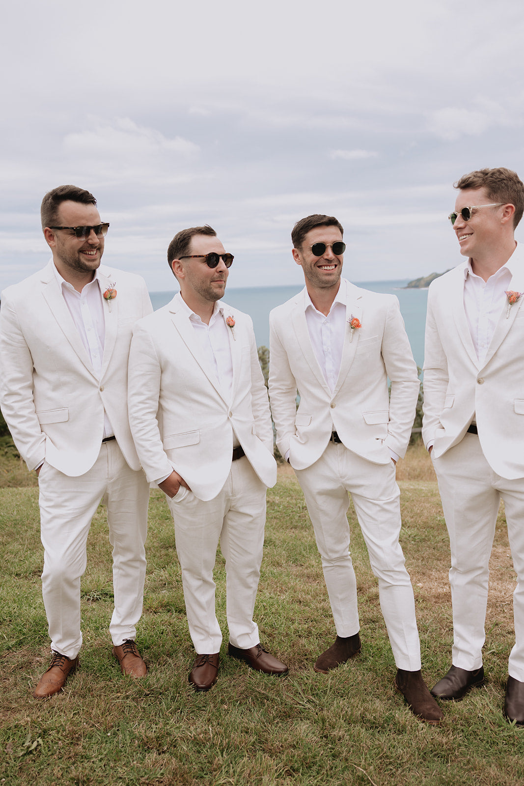 Four men in white suits standing outdoors with a scenic background wearing buttonholes