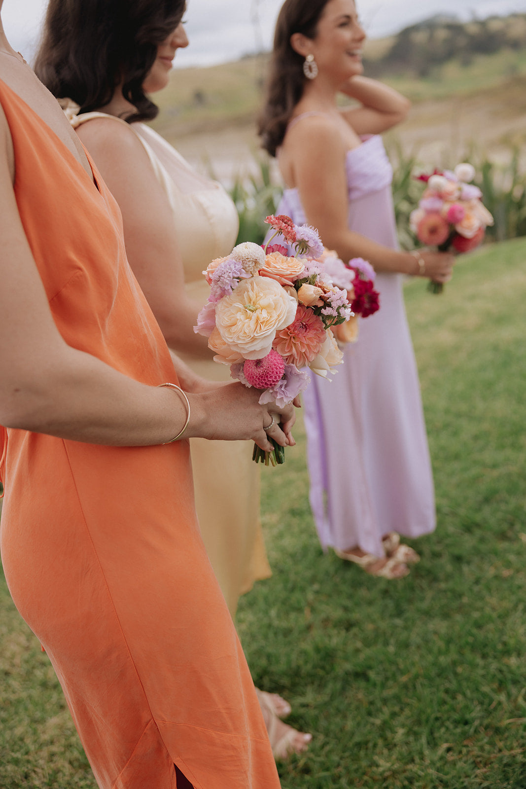 Three women in colourful dresses holding flower bouquets outdoors.