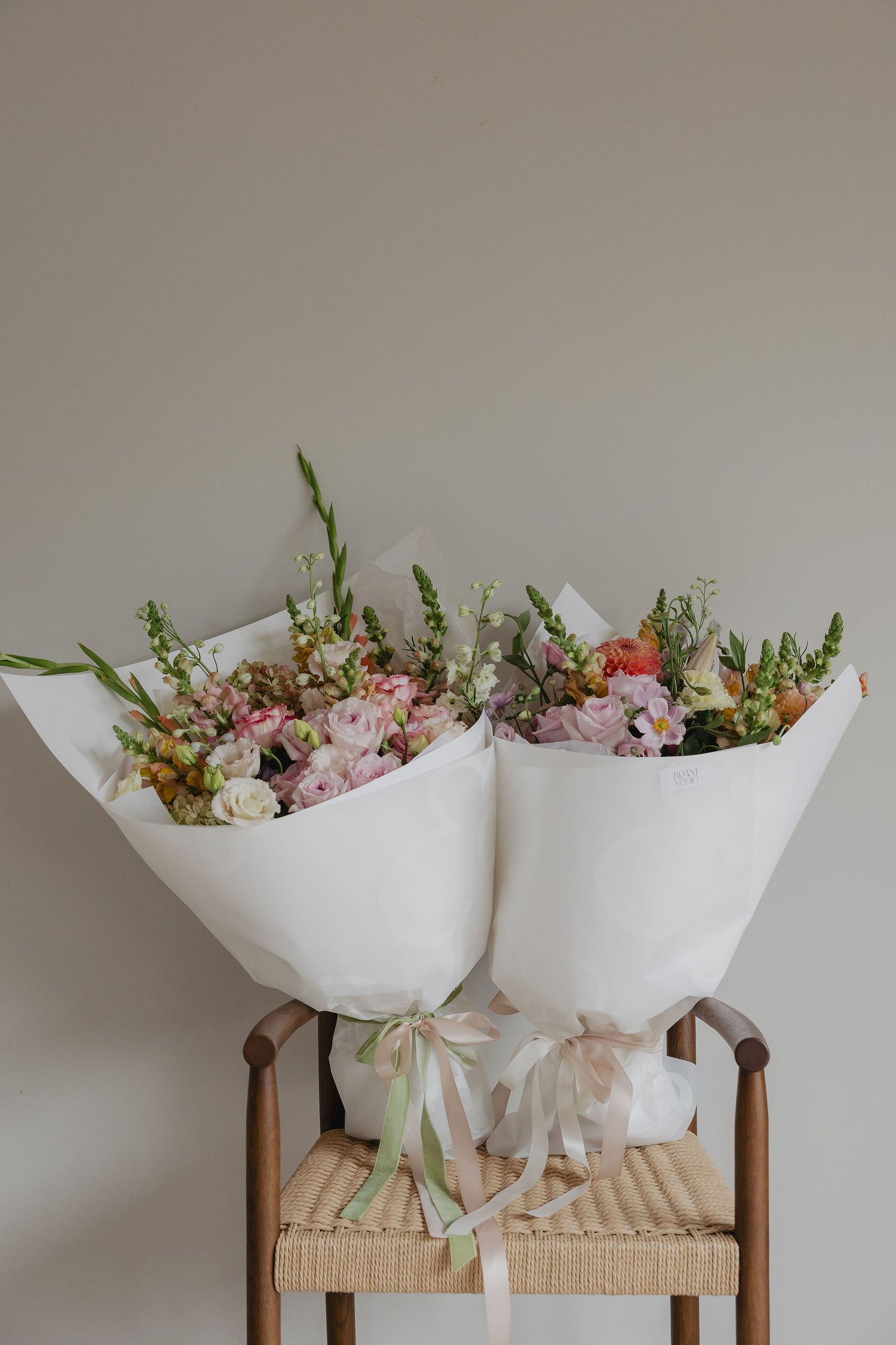 Two flower bouquets wrapped in white paper and tied with ribbons on a wooden chair against a plain wall.