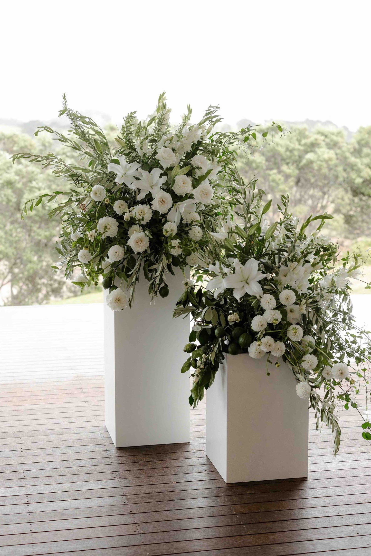 Two white flower arrangements on tall white plinths on a wooden floor with a blurred green outdoor background.