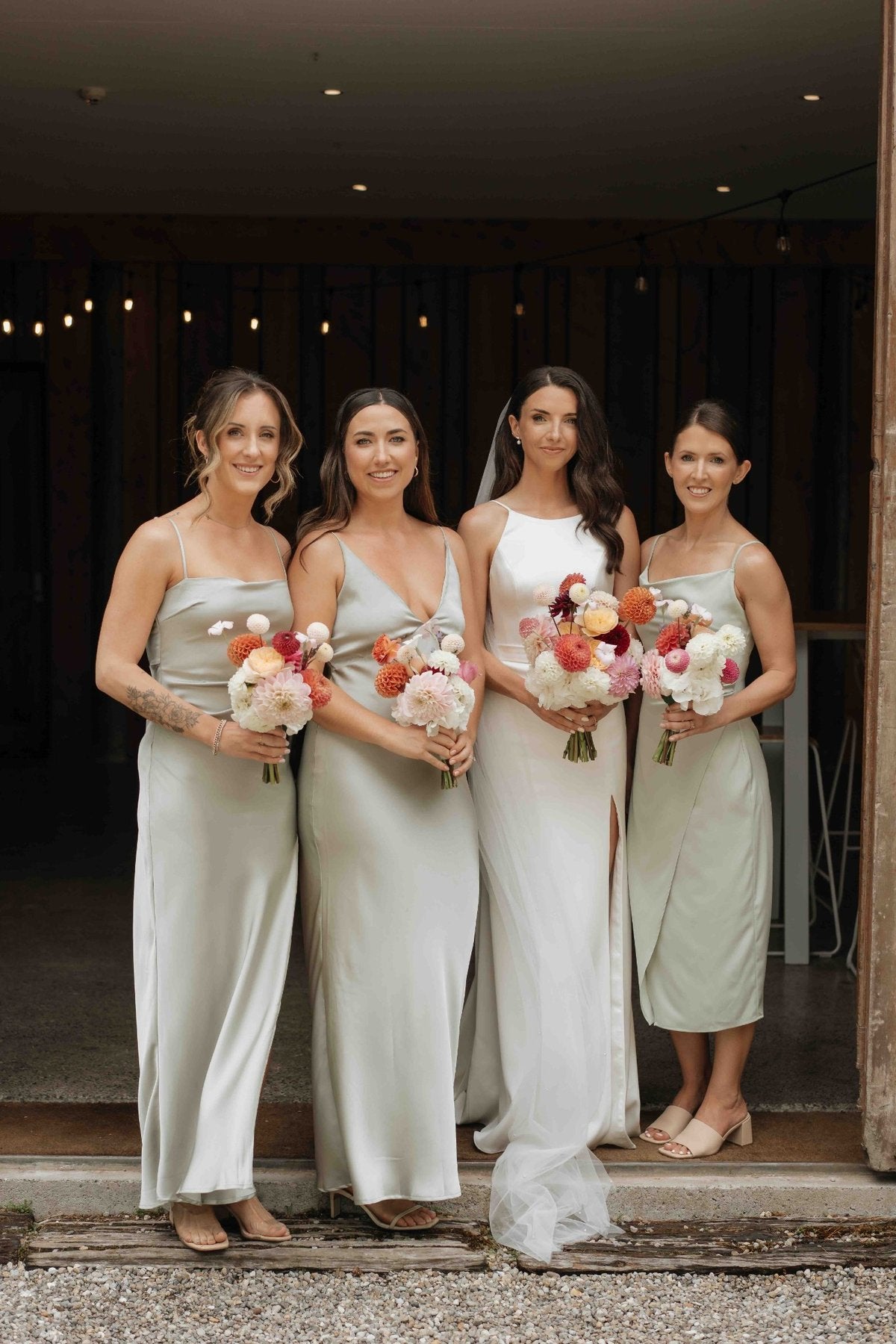 Four women in light-colored dresses holding floral bouquets, standing in a doorway.