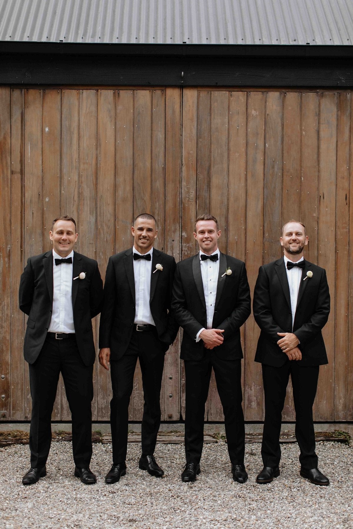 Four men in formal black suits standing in front of a wooden wall.