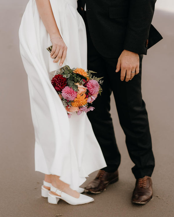 Person in a white dress holding a colorful bouquet, standing next to a person in a black suit on a neutral background.