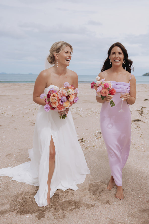 Two women on a beach, one in a white wedding dress and the other in a pink dress, holding floral bouquets. Bride and bridesmaids