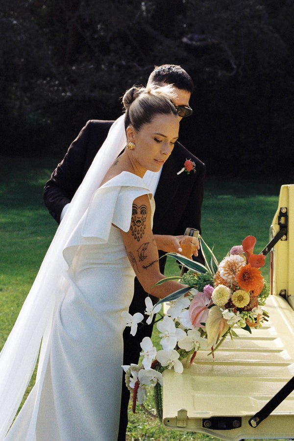 Person in a wedding dress with tattoos holding flowers next to an open casket outdoors.