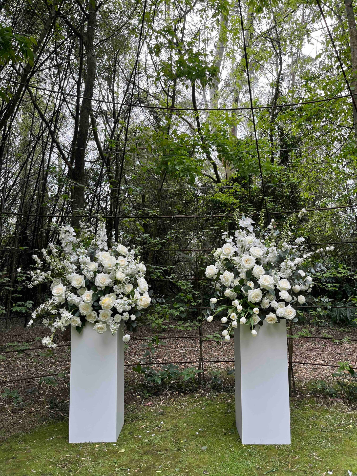 Two white flower wedding plinth arrangements in a forest setting